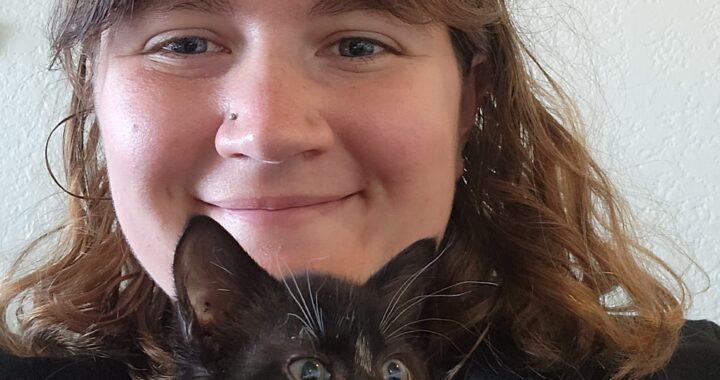 A young woman with bangs smiles at the camera while a small kitten pokes its head out from the top of her hoodie.