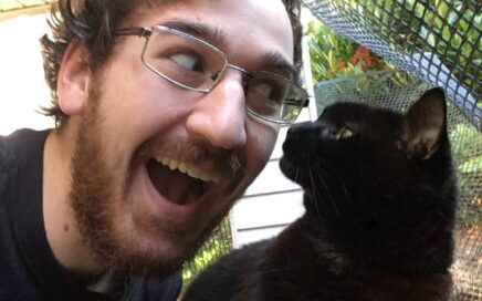 John, our volunteer of the month, leans from left of frame making an exaggeratedly excited expression at a black cat on the right. The cat is very poised and meets his gaze with a mildly curious expression.